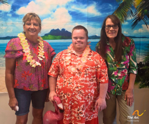 Three adults in bright Hawaiian shirts wearing pose together in front of a tropical beach backdrop; center person holds a pink inflatable flamingo.