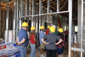 Group of construction workers and visitors wearing yellow hard hats tour an unfinished framing area with metal studs and exposed ceiling joists.